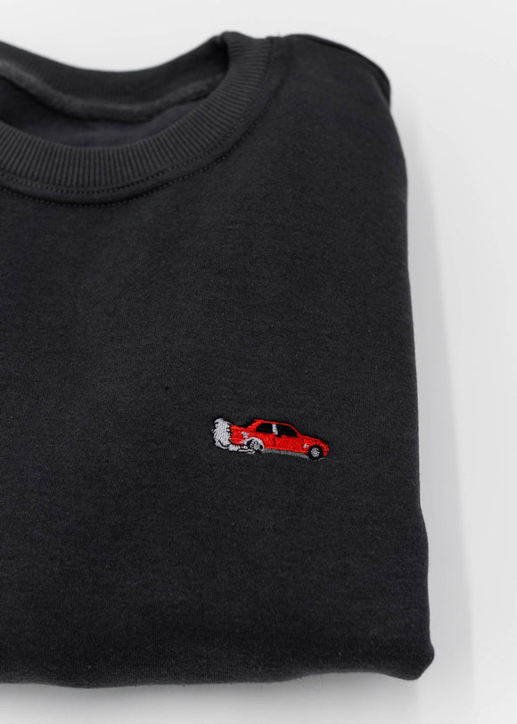 A dark grey BMW/Bimmer E30 M3 crewneck sweater for men. Photo is a close up of the sweater with an embroidered red BMW E30 M3 doing a burnout. Fabric is 100% cotton and high quality and fits to size. The style is long sleeve, crew neck, and embroidery on left chest.
