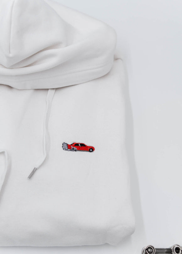 A white BMW/Bimmer unisex hoodie for men and women. Photo is a close up of the sweater with an embroidered Red BMW E30 M3 doing a burnout. Fabric composition is cotton, polyester, and rayon. The material is very soft, stretchy, and non-transparent. The style of this hoodie is long sleeve, crewneck with a hood, hooded, with embroidery on the left chest.