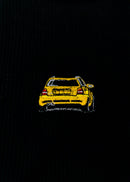 A black Audi crop top for women. Photo is a close up view of the top with an embroidered imola yellow Audi B5 RS4. Fabric composition is polyester, and elastine. The material is stretchy, ribbed, and non-transparent. The style of this shirt is sleeveless, with a crewneck neckline.