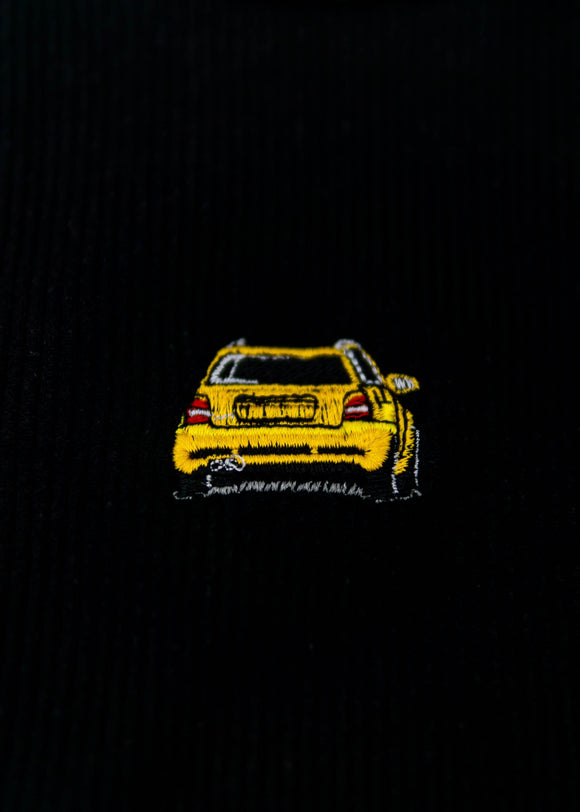 A black Audi crop top for women. Photo is a close up view of the top with an embroidered imola yellow Audi B5 RS4. Fabric composition is polyester, and elastine. The material is stretchy, ribbed, and non-transparent. The style of this shirt is sleeveless, with a crewneck neckline.