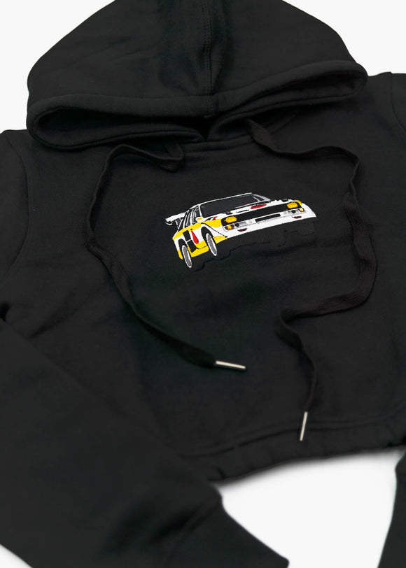 A black Audi cropped hoodie for women. Photo is a front view of the cropped sweater with an embroidered Sport Quattro S1 E2. Fabric composition is cotton, and polyester. The material is soft, stretchy, and non-transparent. The style of this crop hoodie is long sleeve, crewneck with a hood, hooded, with embroidery on the chest.