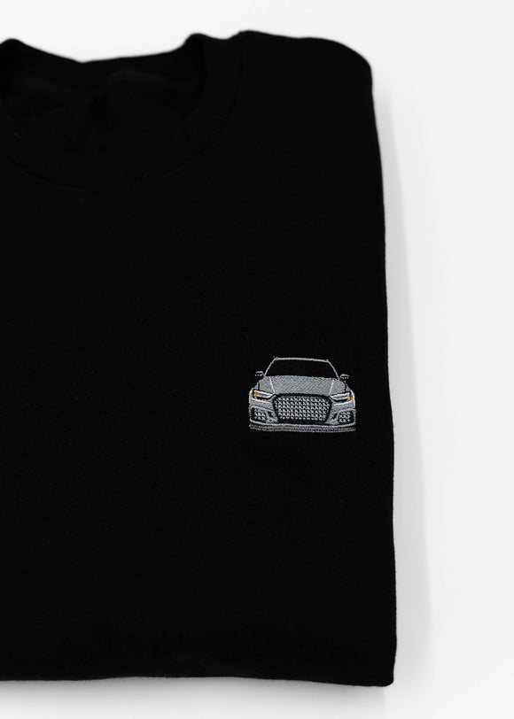 Close up of an embroidered 8V RS3 on a black men's high quality crewneck sweater. Photo shows the high quality detailed embroidery of a nardo grey 8V RS3. Fabric composition of the shirt is cotton and polyester. The material is very soft, stretchy, and non-transparent. The style of this sweater is a crewneck, long sleeve, elastic bottom, with embroidery on the left chest.