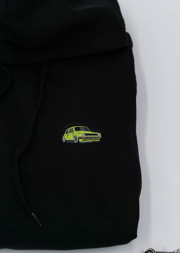 A black VW Volkswagen unisex hoodie for men and women. Photo is a close up of the sweater with an embroidered modified green Mk1 Golf. Fabric composition is cotton, polyester, and rayon. The material is very soft, stretchy, and non-transparent. The style of this hoodie is long sleeve, crewneck with a hood, hooded, with embroidery on the left chest.