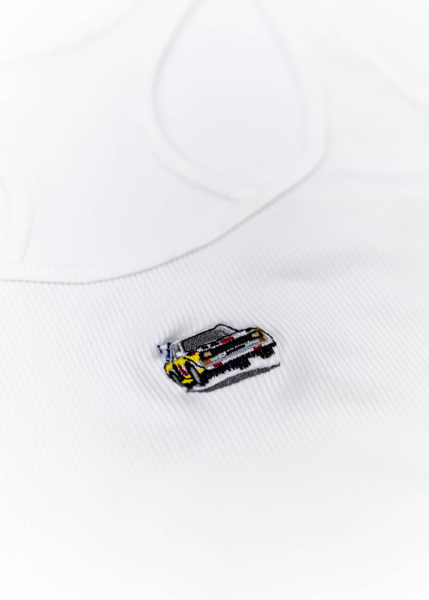A white Audi crop top for women. Photo is a close up view of the top with an embroidered Audi Sport Quattro S1 E2. Fabric composition is polyester, and cotton. The material is stretchy, ribbed, and non-transparent. The style of this shirt is sleeveless, with a scoop neckline.