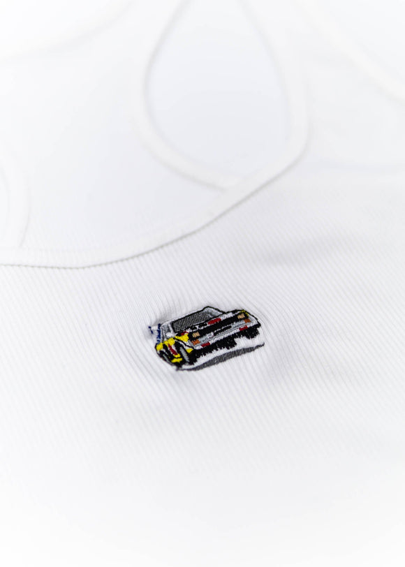 A white Audi crop top for women. Photo is a close up view of the top with an embroidered Audi Sport Quattro S1 E2. Fabric composition is polyester, and cotton. The material is stretchy, ribbed, and non-transparent. The style of this shirt is sleeveless, with a scoop neckline.