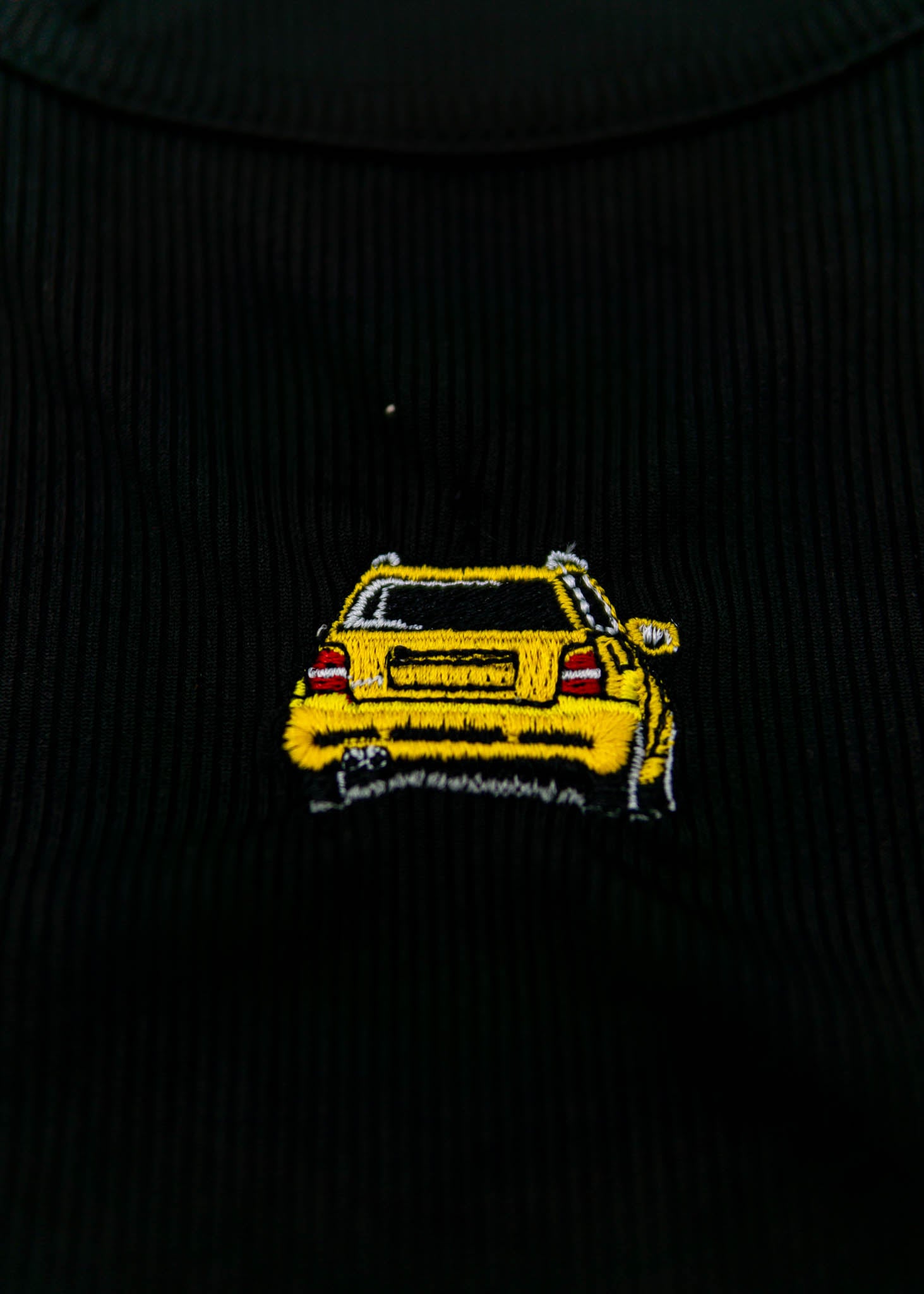 A black Audi crop top for women. Photo is a close up view of the top with an embroidered imola yellow Audi B5 RS4. Fabric composition is polyester, and elastine. The material is stretchy, ribbed, and non-transparent. The style of this shirt is sleeveless, with a crewneck neckline.