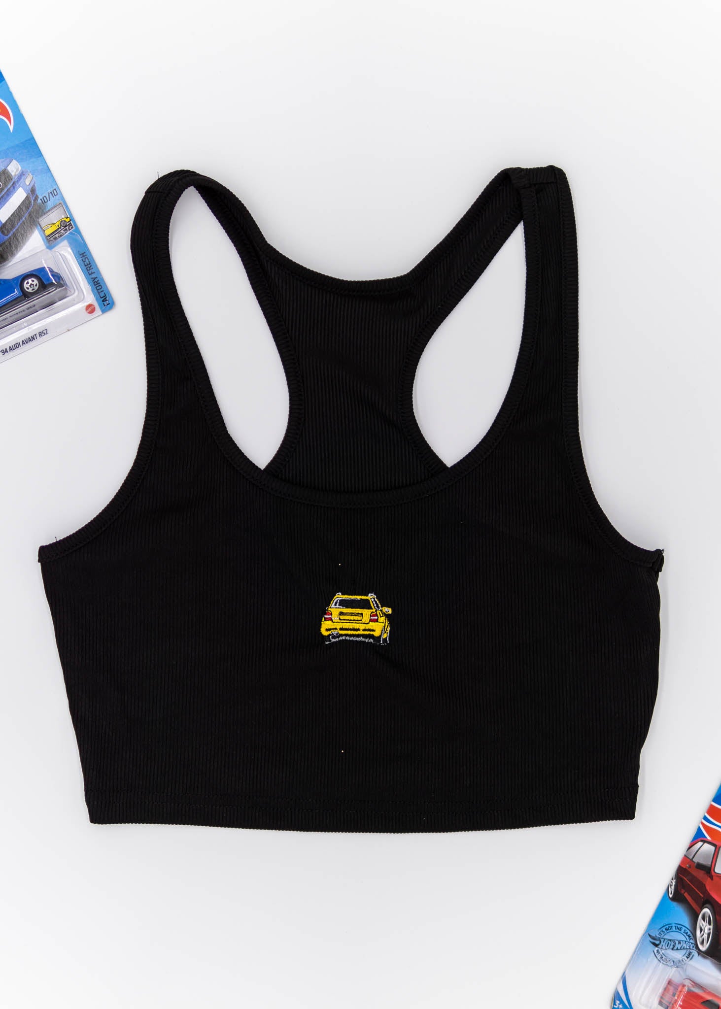 A black Audi crop top for women. Photo is a front view of the top with an embroidered imola yellow Audi B5 RS4. Fabric composition is polyester, and elastine. The material is stretchy, ribbed, and non-transparent. The style of this shirt is sleeveless, with a crewneck neckline.