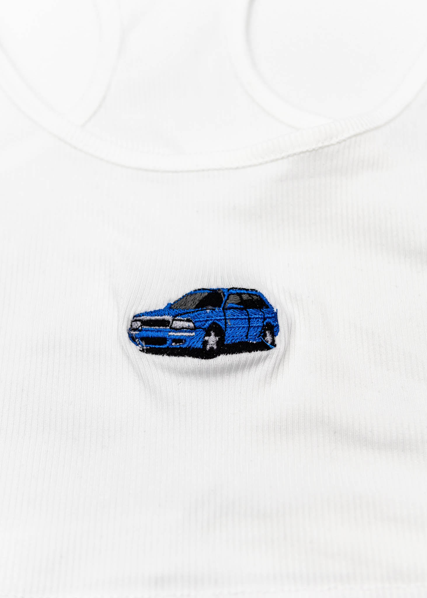A white Audi crop top for women. Photo is a close up view of the top with an embroidered Nogaro Blue Audi 80 RS2 Avant made by Porsche. Fabric composition is polyester, and cotton. The material is stretchy, ribbed, and non-transparent. The style of this shirt is sleeveless, with a scoop neckline.