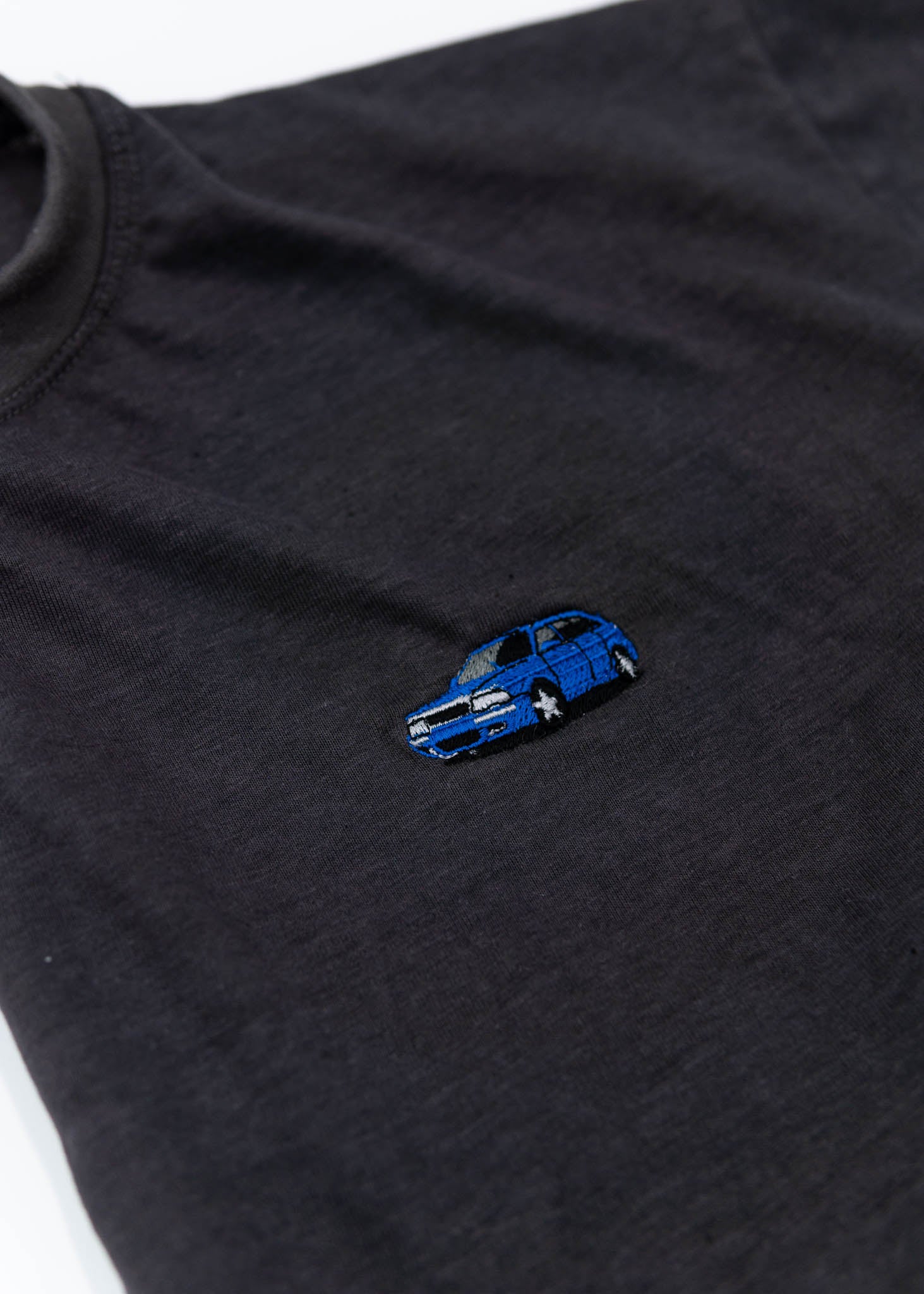 A dark grey cropped t-shirt for Audi women. Close up view of a 100% cotton crop tee with an embroidered Nogaro Blue Audi 80 RS2 Avant made by Porsche. The material is stretchy, and non-transparent with a crewneck neckline and short sleeves.