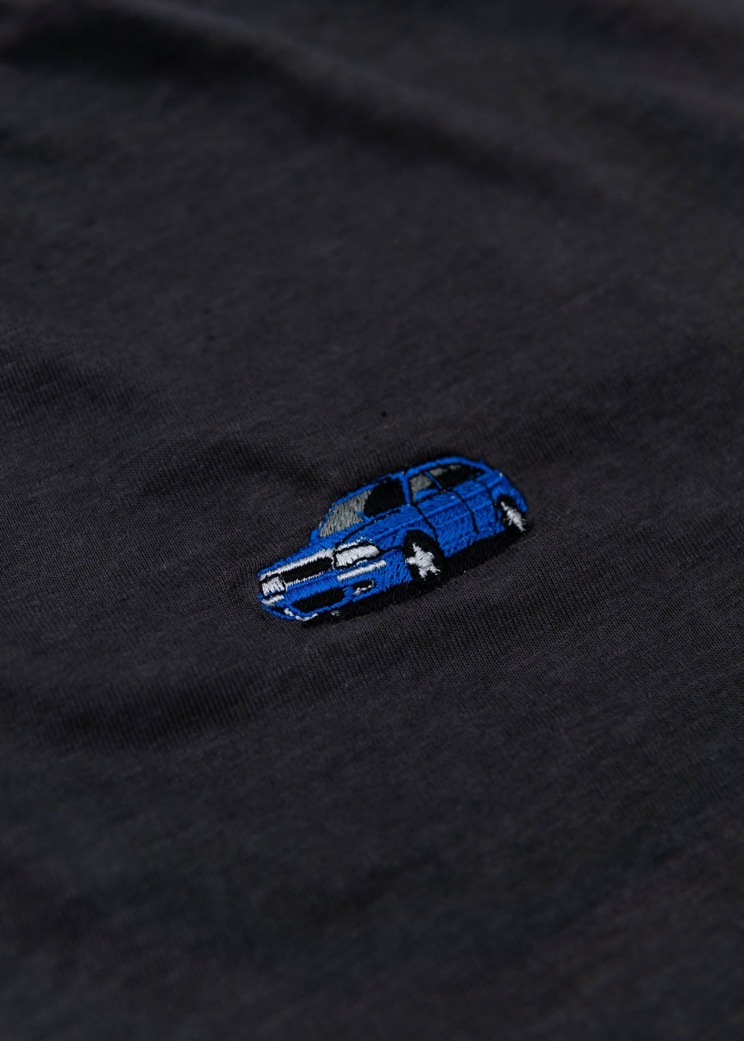 A dark grey cropped t-shirt for Audi women. Close up view of a 100% cotton crop tee with an embroidered Nogaro Blue Audi 80 RS2 Avant made by Porsche. The material is stretchy, and non-transparent with a crewneck neckline and short sleeves.