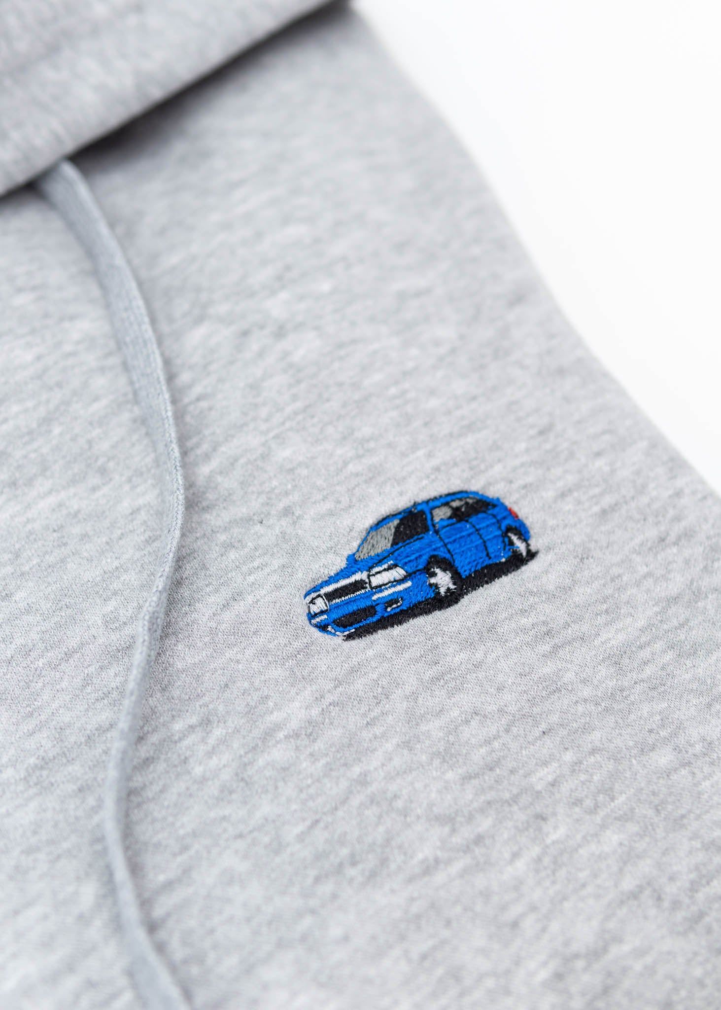 A grey Audi unisex hoodie for men and women. Photo is a close up view of the sweater with an embroidered Nogaro Blue Audi 80 RS2 Avant made by Porsche. Fabric composition is cotton, polyester, and rayon. The material is very soft, stretchy, and non-transparent. The style of this hoodie is long sleeve, crewneck with a hood, hooded, with embroidery on the left chest.