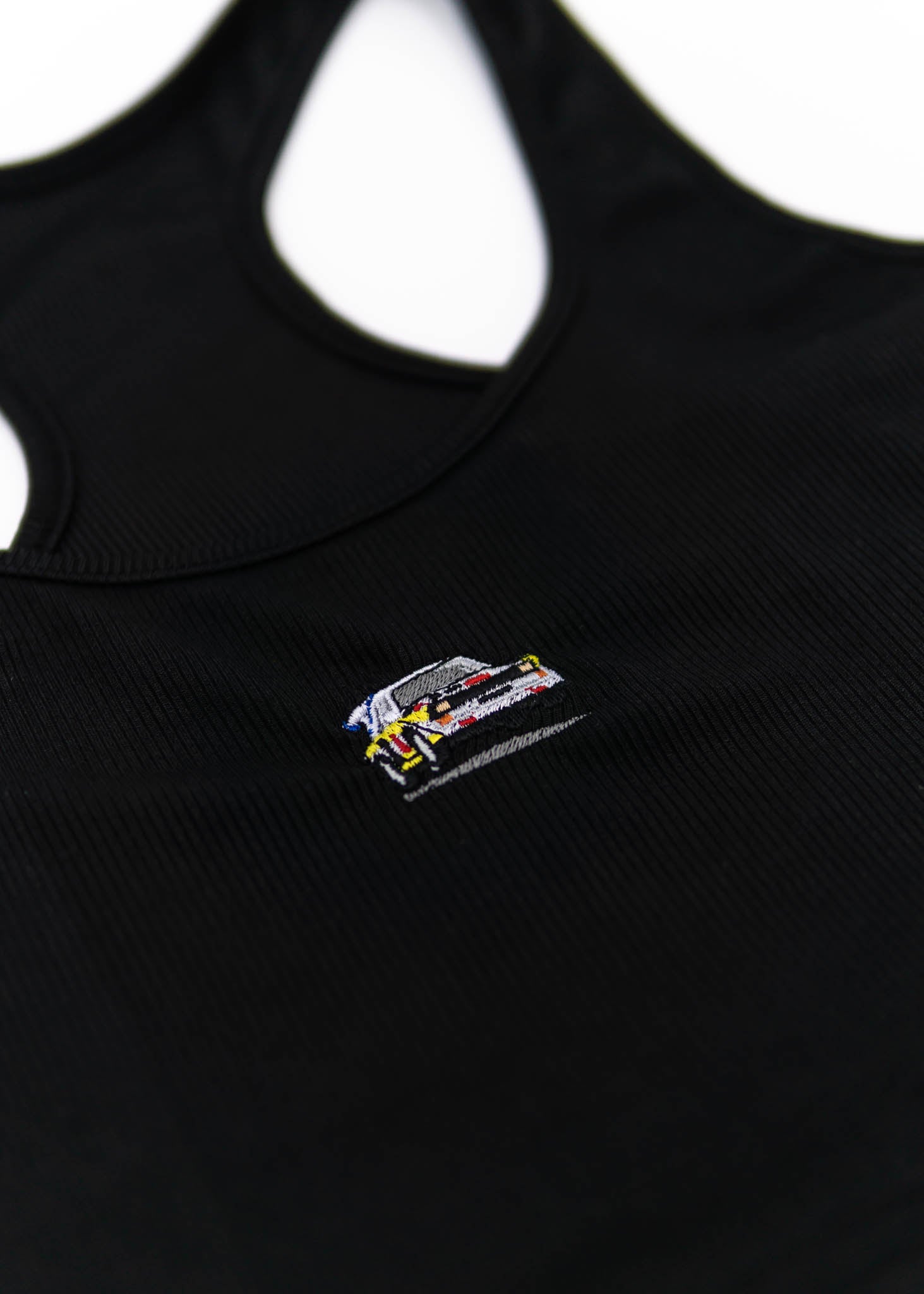 A black Audi crop top for women. Photo is a close up view of the top with an embroidered Audi Sport Quattro S1 E2. Fabric composition is polyester, and cotton. The material is stretchy, ribbed, and non-transparent. The style of this shirt is sleeveless, with a scoop neckline.