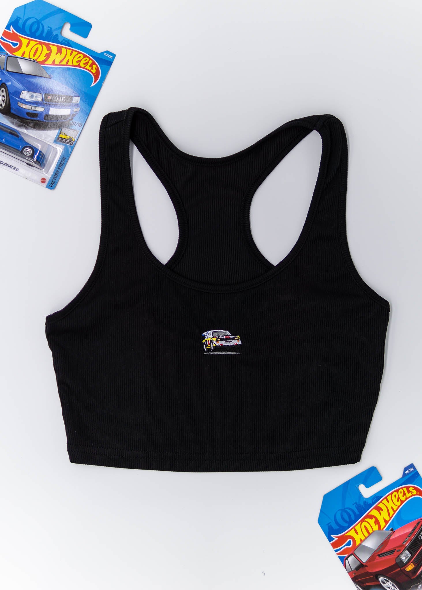 A black Audi crop top for women. Photo is a front view of the top with an embroidered Audi Sport Quattro S1 E2. Fabric composition is polyester, and cotton. The material is stretchy, ribbed, and non-transparent. The style of this shirt is sleeveless, with a scoop neckline.