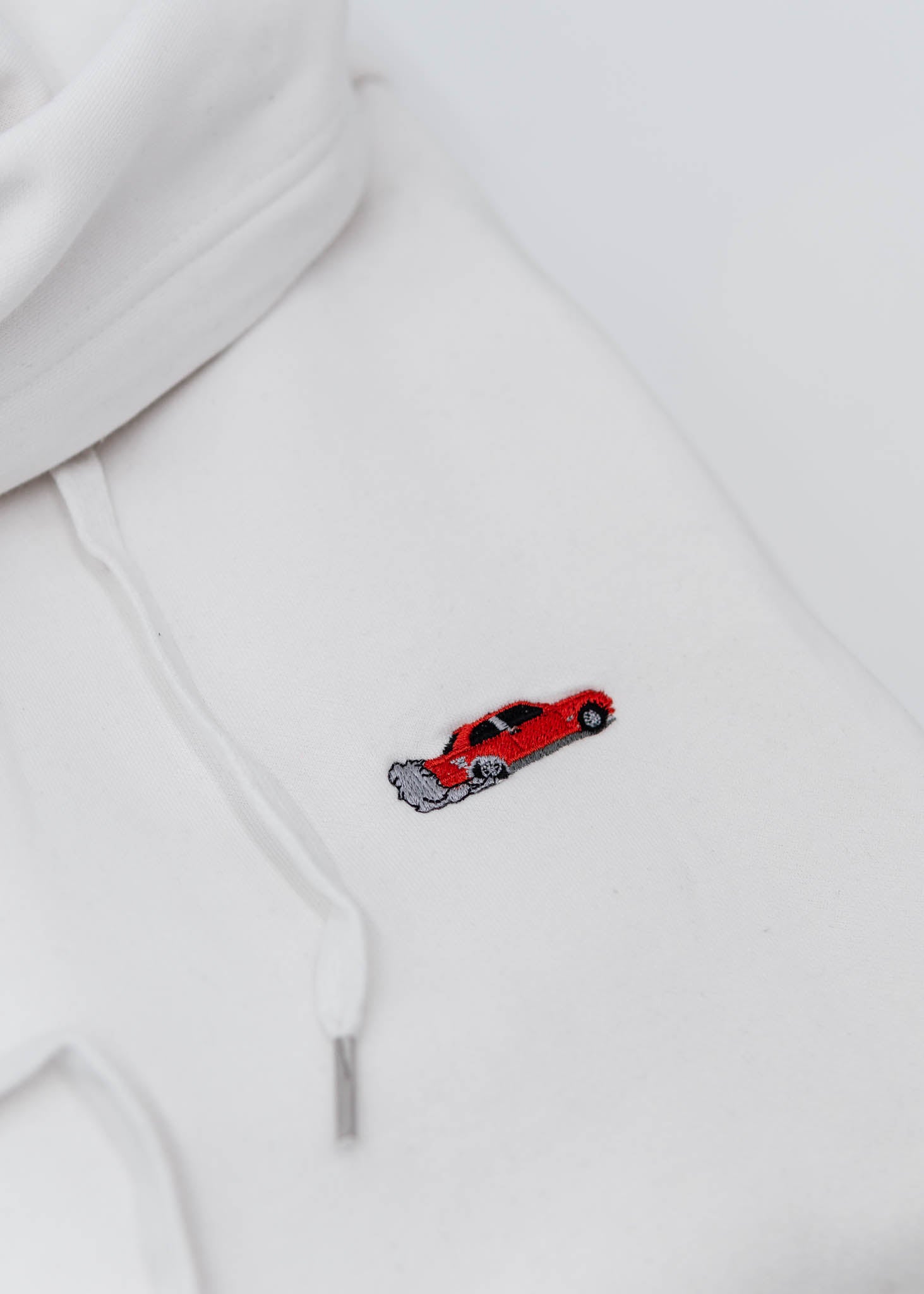 A white BMW/Bimmer unisex hoodie for men and women. Photo is a close up of the sweater with an embroidered Red BMW E30 M3 doing a burnout. Fabric composition is cotton, polyester, and rayon. The material is very soft, stretchy, and non-transparent. The style of this hoodie is long sleeve, crewneck with a hood, hooded, with embroidery on the left chest.