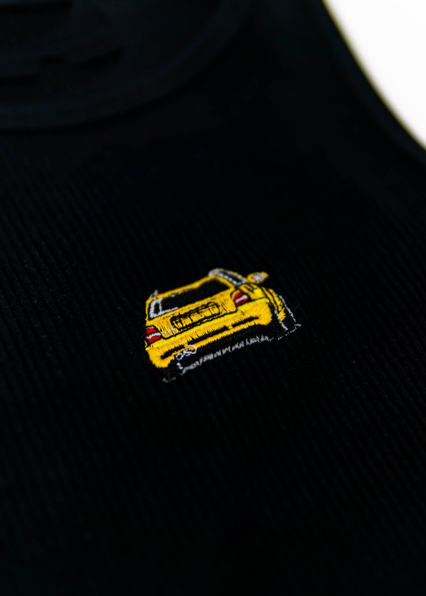 A black Audi crop top for women. Photo is a close up view of the top with an embroidered imola yellow Audi B5 RS4. Fabric composition is polyester, and elastine. The material is stretchy, ribbed, and non-transparent. The style of this shirt is sleeveless, with a crewneck neckline.