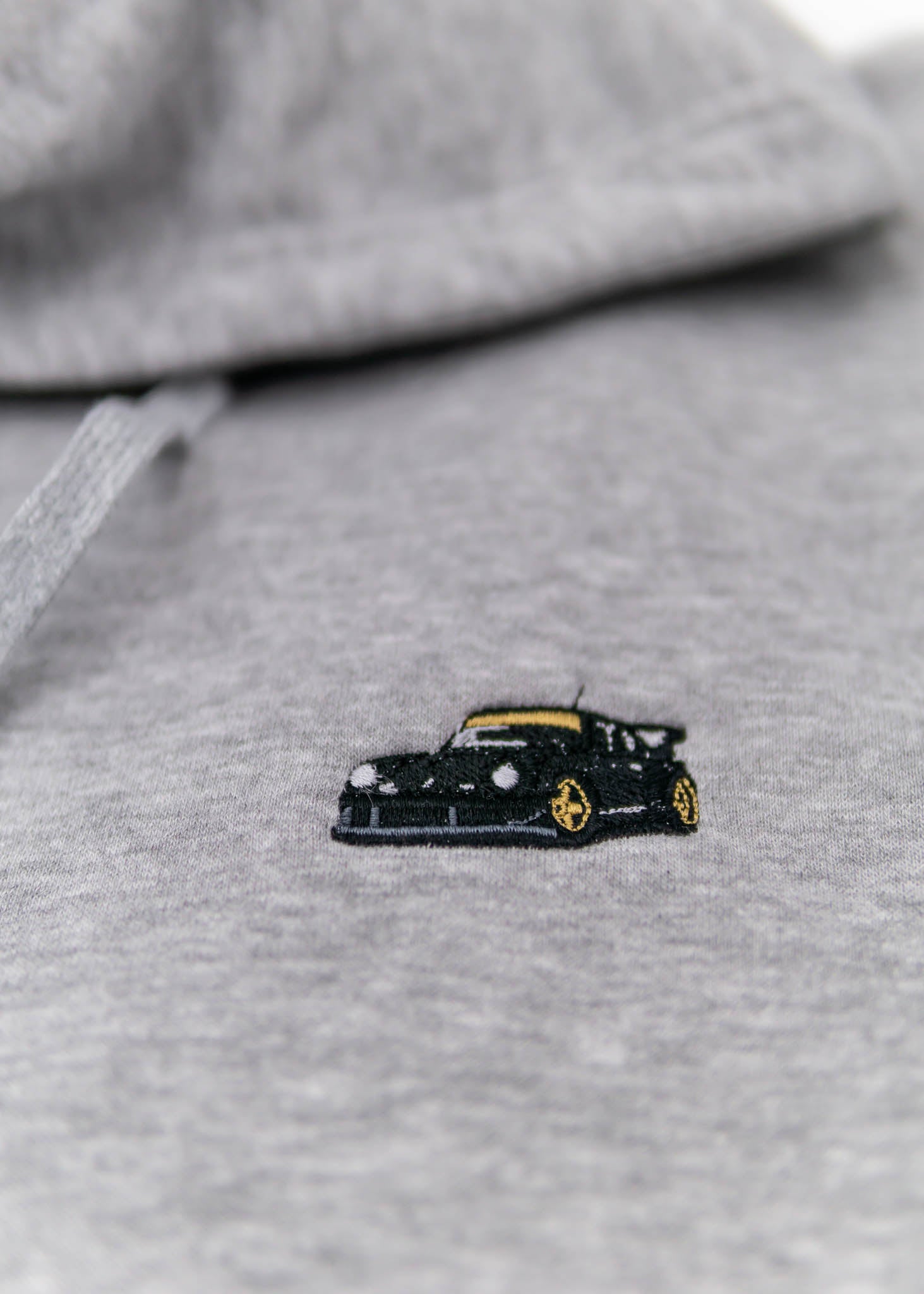 A grey Porsche unisex hoodie for men and women. Photo is a close up view of the sweater with an embroidered RWB Porsche 930 911 Turbo Akira Nakai Stella Artois. Fabric composition is cotton, polyester, and rayon. The material is very soft, stretchy, and non-transparent. The style of this hoodie is long sleeve, crewneck with a hood, hooded, with embroidery on the left chest.