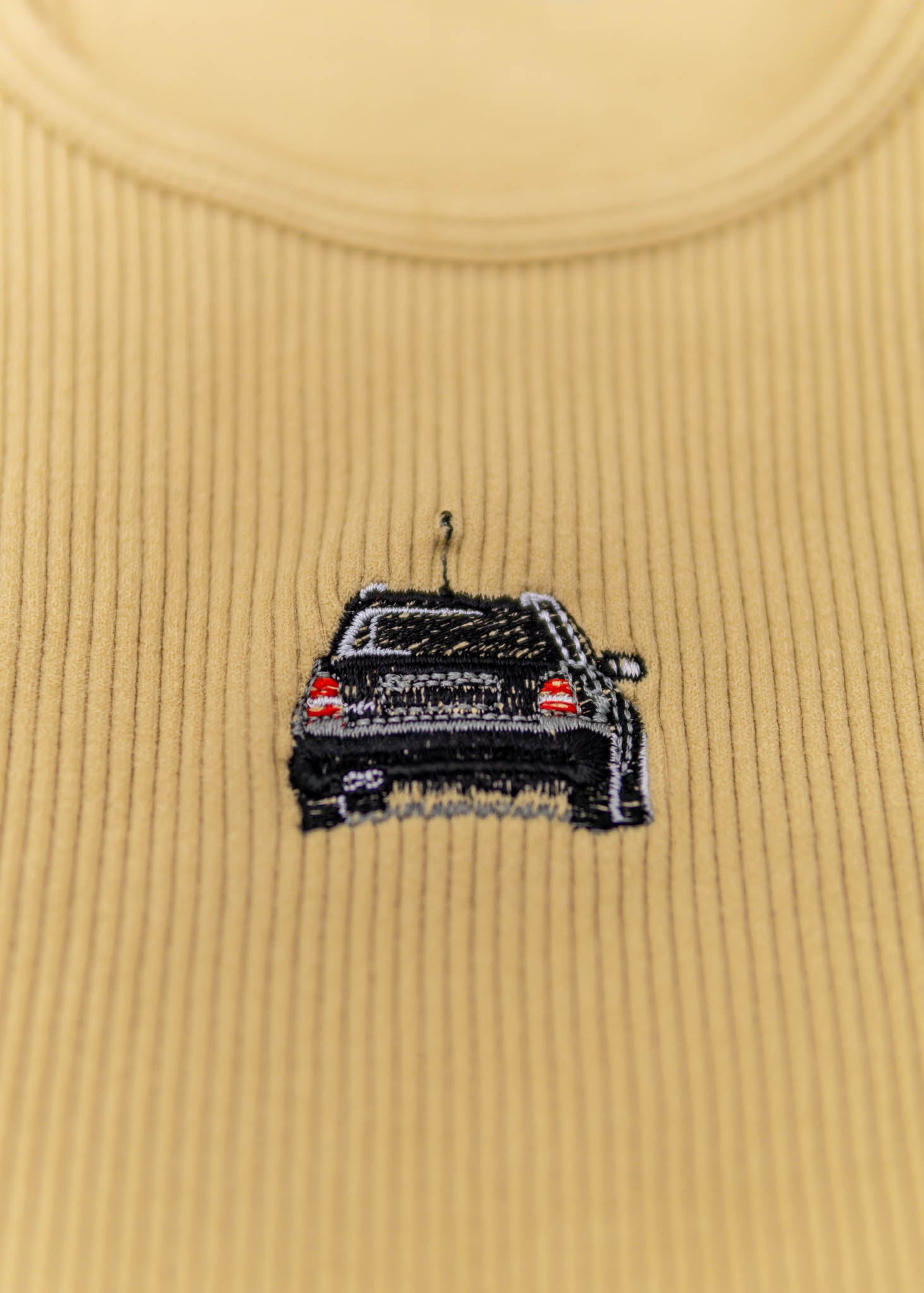 A khaki Audi crop top for women. Photo is a close up view of the top with an embroidered black Audi B5 RS4. Fabric composition is polyester, and elastine. The material is stretchy, ribbed, and non-transparent. The style of this shirt is sleeveless, with a crewneck neckline.