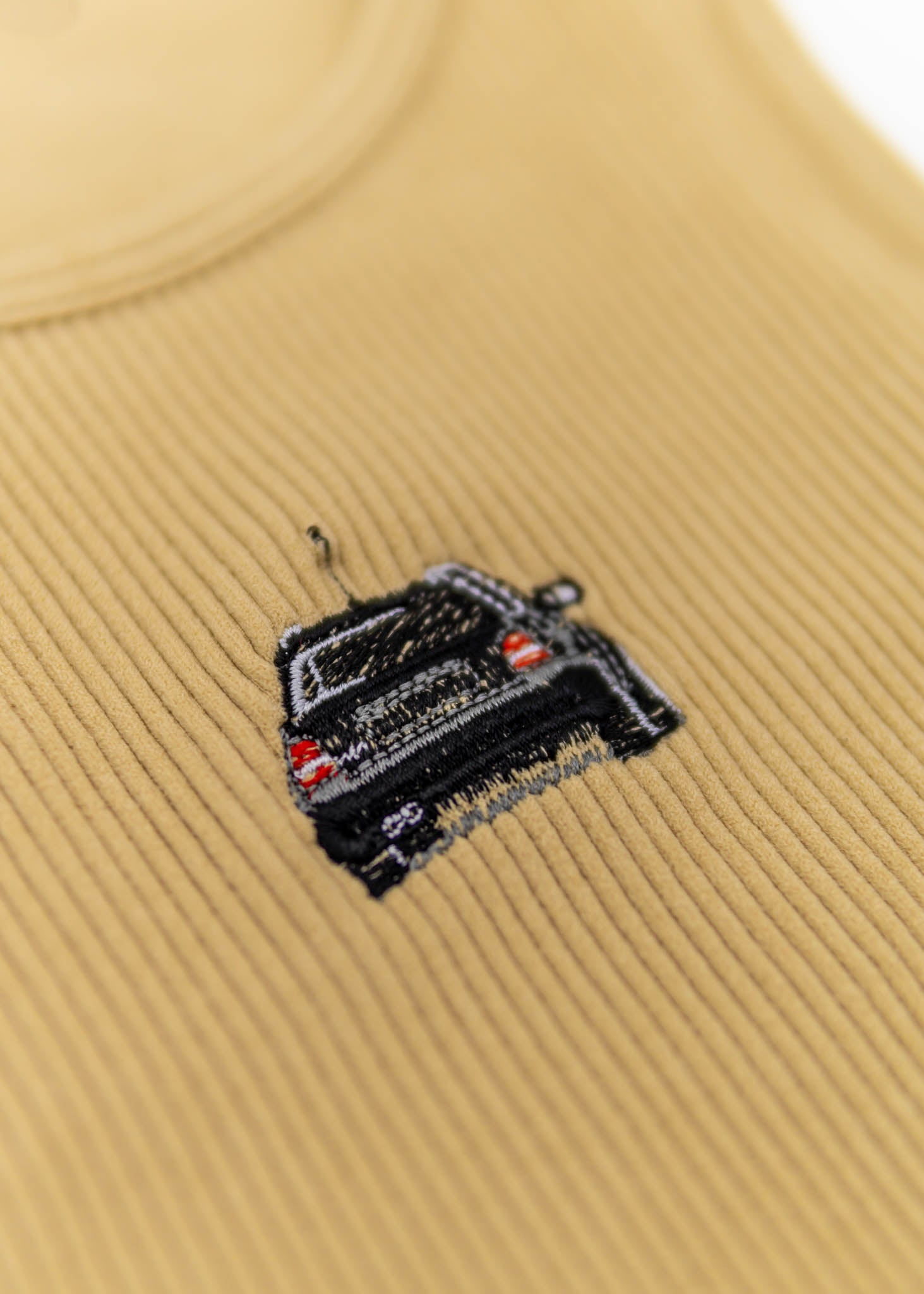 A khaki Audi crop top for women. Photo is a close up view of the top with an embroidered black Audi B5 RS4. Fabric composition is polyester, and elastine. The material is stretchy, ribbed, and non-transparent. The style of this shirt is sleeveless, with a crewneck neckline.