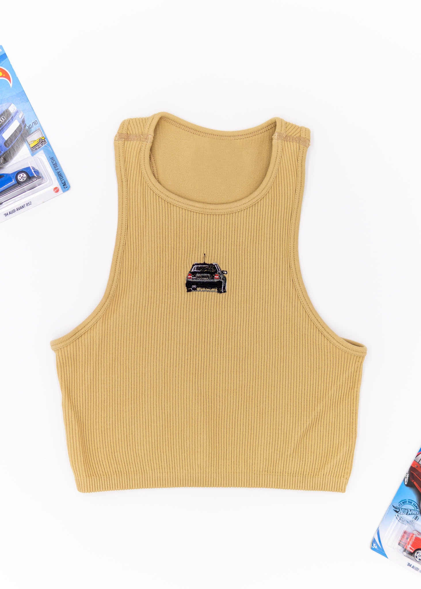 A khaki Audi crop top for women. Photo is a front view of the top with an embroidered black Audi B5 RS4. Fabric composition is polyester, and elastine. The material is stretchy, ribbed, and non-transparent. The style of this shirt is sleeveless, with a crewneck neckline.