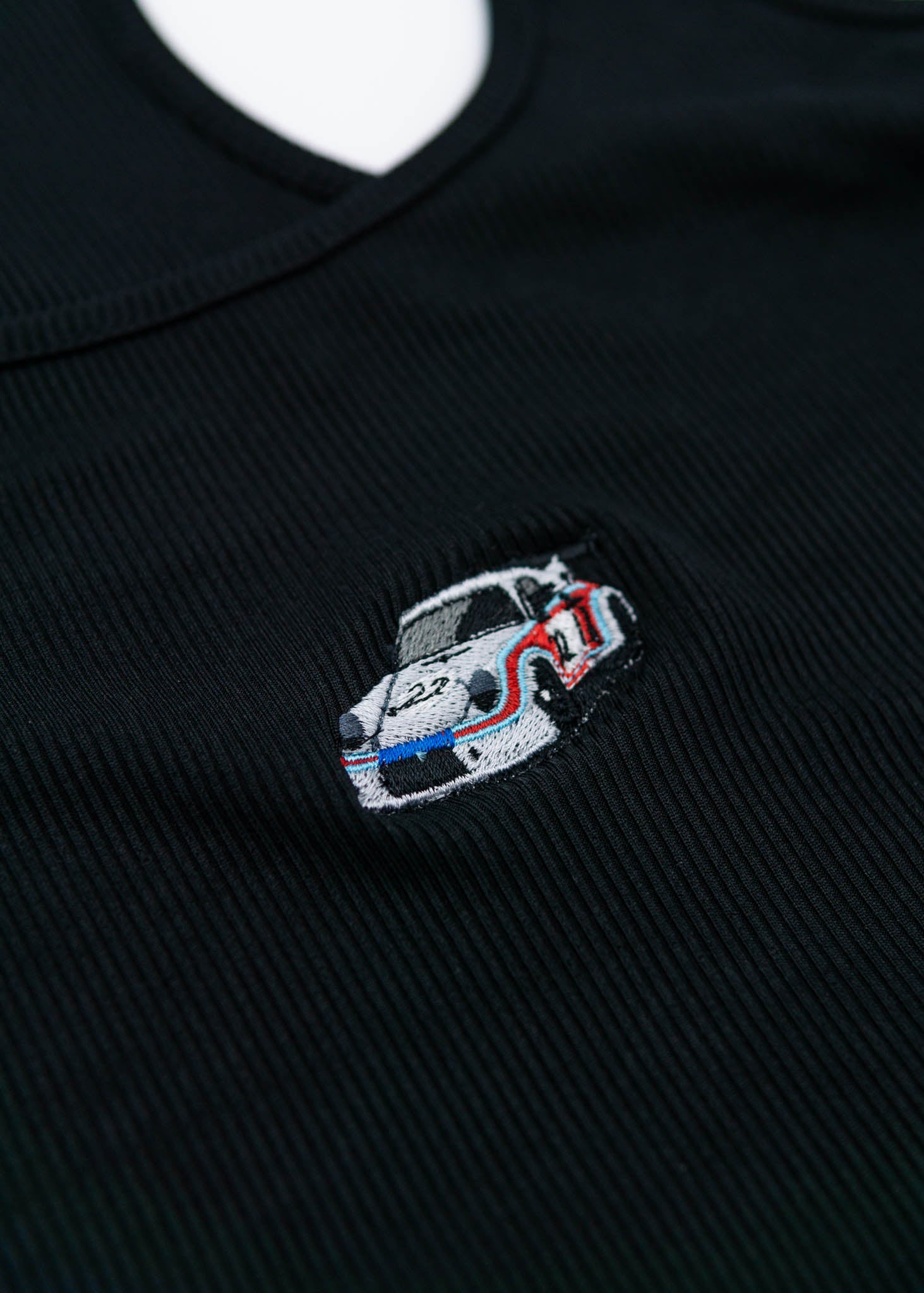 A black Porsche crop top for women. Photo is a close up of the top with an embroidered Martini R13 Porsche 911 Carrera RSR 2.1 Turbo. Fabric composition is polyester, and cotton. The material is stretchy, ribbed, and non-transparent. The style of this shirt is sleeveless, with a scoop neckline.