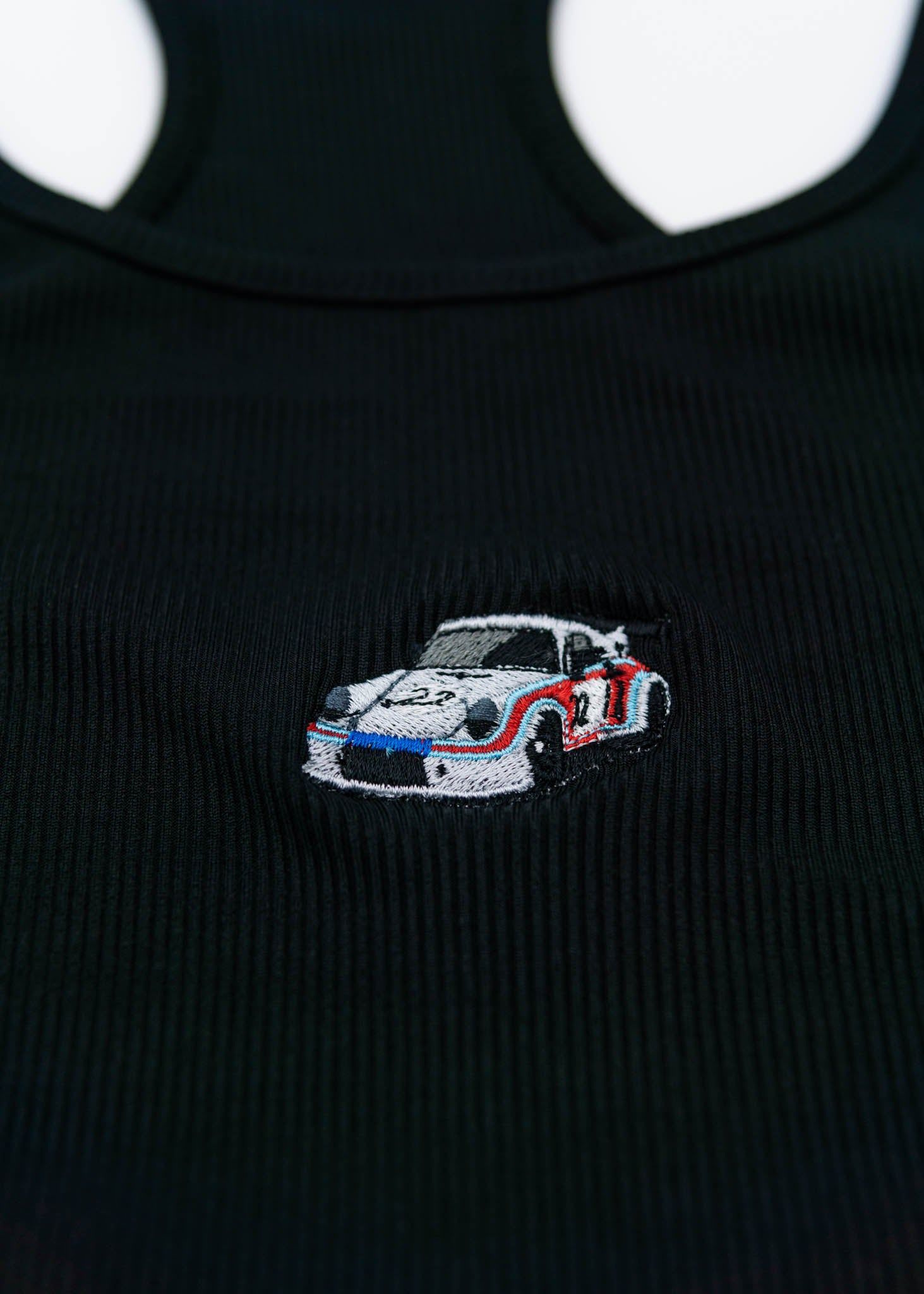 A black Porsche crop top for women. Photo is a close up of the top with an embroidered Martini R13 Porsche 911 Carrera RSR 2.1 Turbo. Fabric composition is polyester, and cotton. The material is stretchy, ribbed, and non-transparent. The style of this shirt is sleeveless, with a scoop neckline.