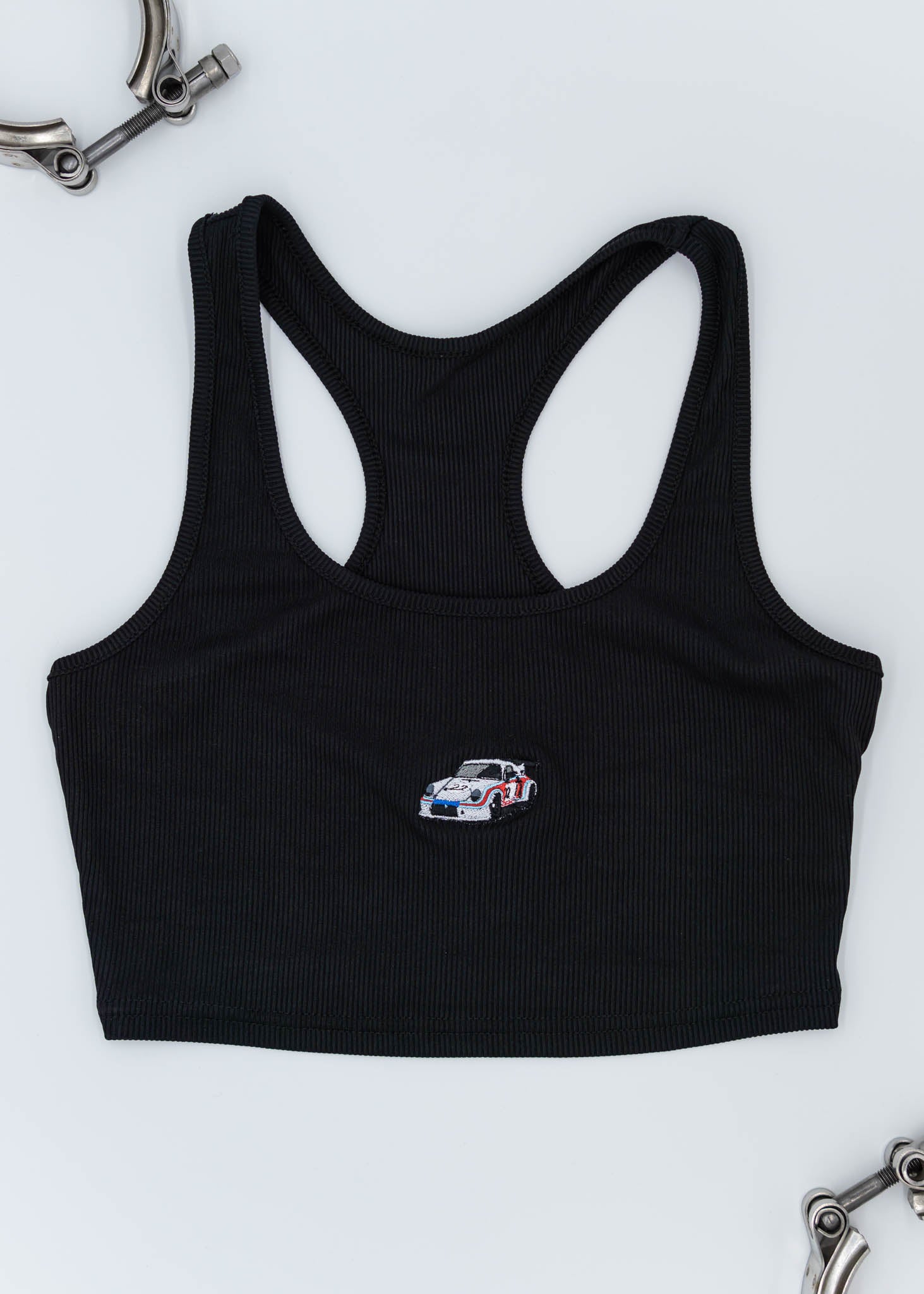 A black Porsche crop top for women. Photo is the front of the top with an embroidered Martini R13 Porsche 911 Carrera RSR 2.1 Turbo. Fabric composition is polyester, and cotton. The material is stretchy, ribbed, and non-transparent. The style of this shirt is sleeveless, with a scoop neckline.