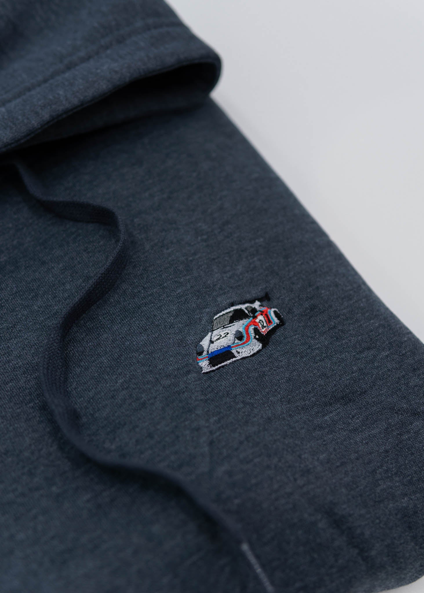 A dark grey Porsche racing unisex hoodie for men and women. Photo is a close up of the sweater with an embroidered Martini R13 Porsche 911 Carrera RSR 2.1 Turbo. Fabric composition is cotton, polyester, and rayon. The material is very soft, stretchy, and non-transparent. The style of this hoodie is long sleeve, crewneck with a hood, hooded, with embroidery on the left chest.