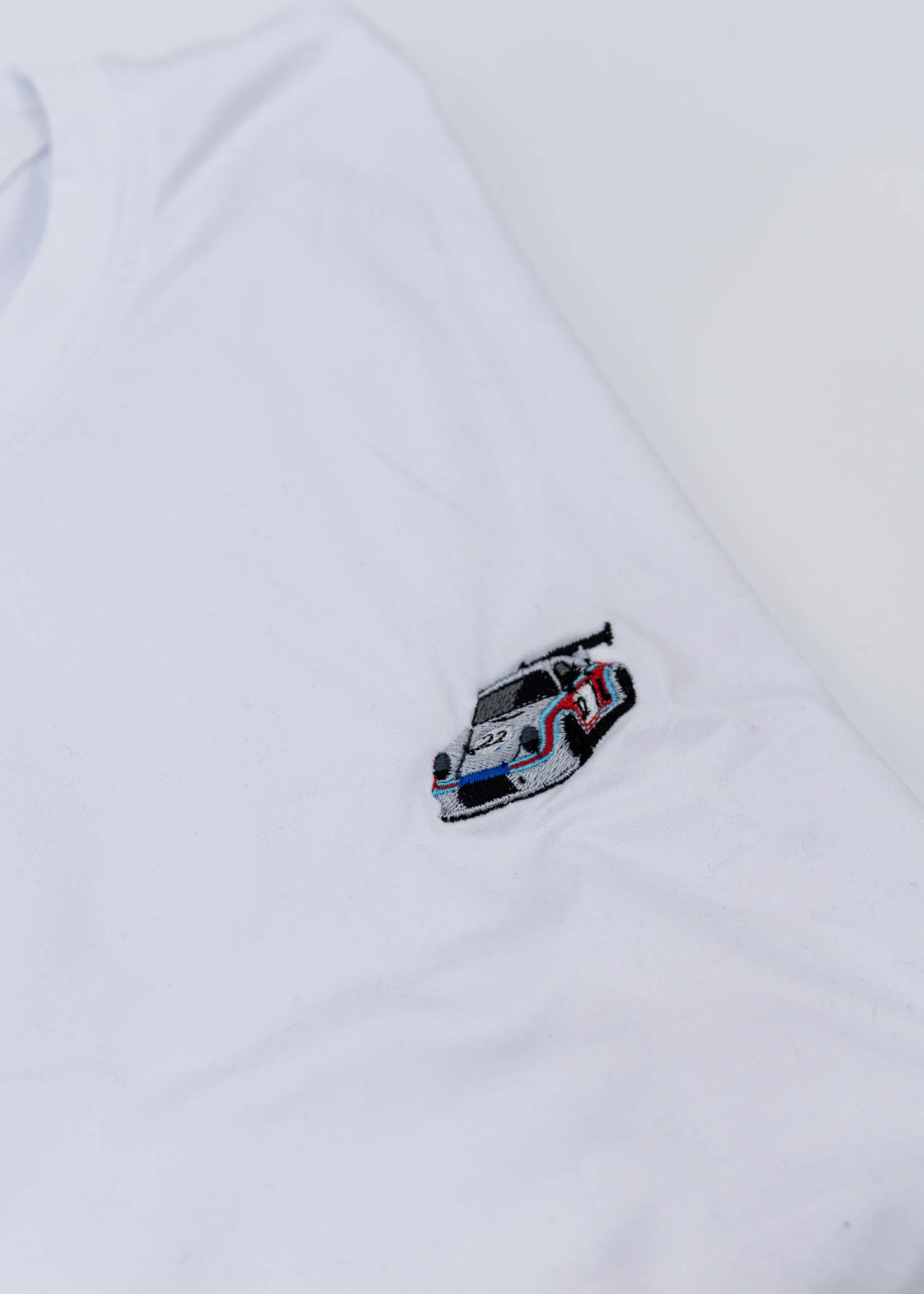 A white Porsche T-Shirt for men. Photo is a close up of the shirt with an embroidered Martini R13 Porsche 911 Carrera RSR 2.1 Turbo. Fabric composition is 100% polyester. The material is very stretchy, soft, comfortable, breathable, and non-transparent. The style of this shirt is short sleeve, with a crewneck neckline.