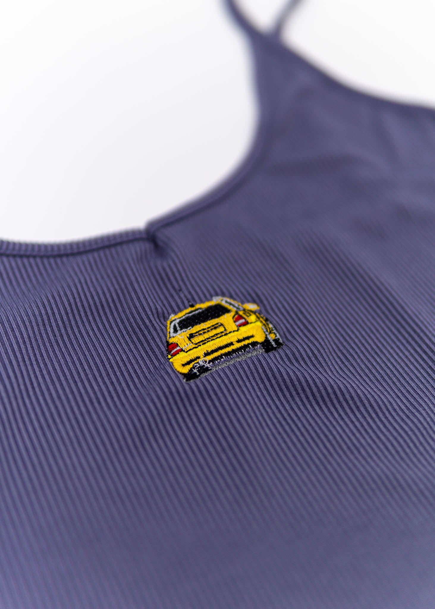 A purple Audi crop top for plus size women. Photo is a close up view of the top with an embroidered Imola Yellow Audi B5 RS4. Fabric composition is polyester, and spandex. The material is stretchy, ribbed, and non-transparent. The style of this shirt is sleeveless, with a deep neckline.