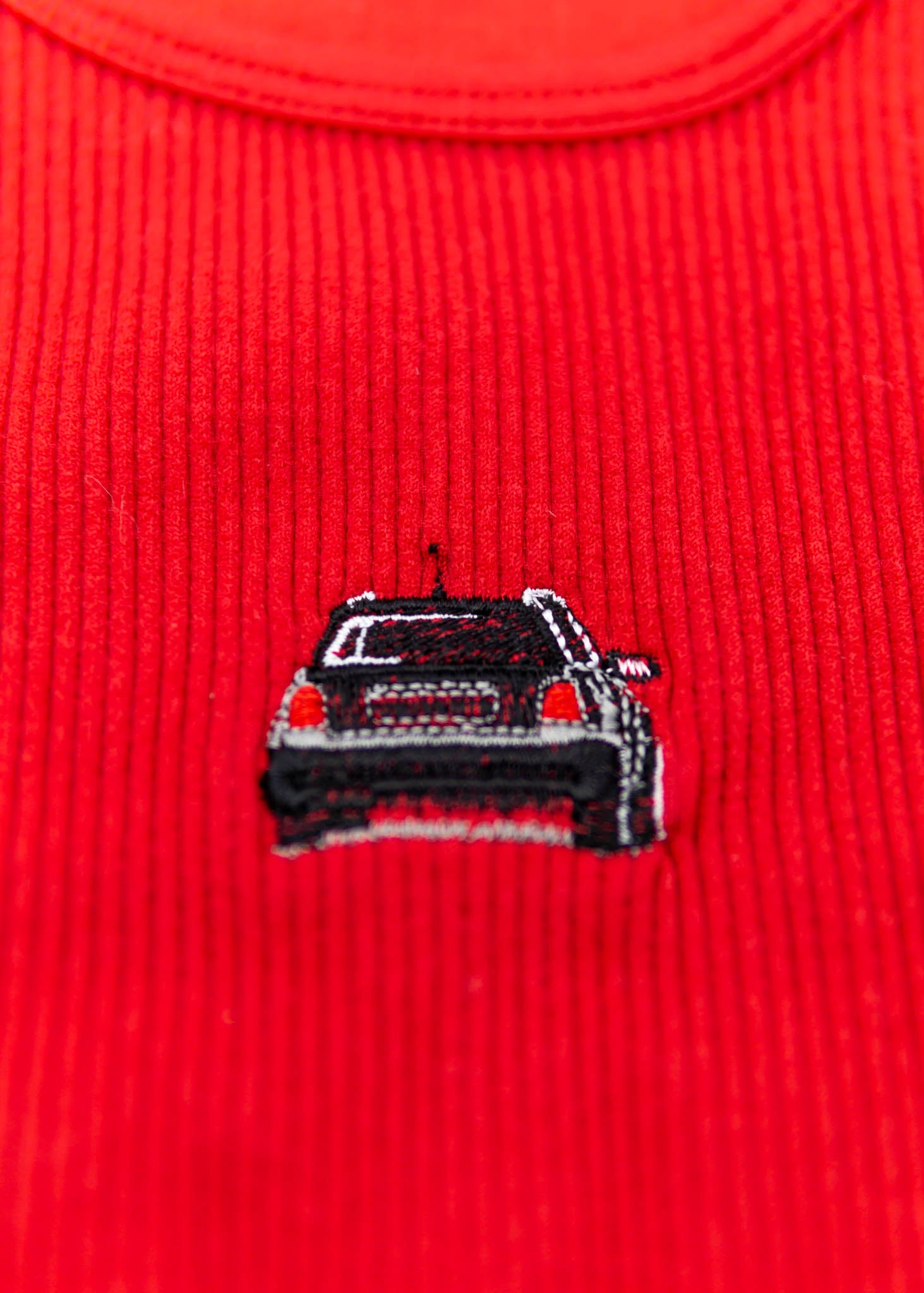 A red Audi crop top for women. Photo is a close up view of the top with an embroidered black Audi B5 RS4. Fabric composition is polyester, and elastine. The material is stretchy, ribbed, and non-transparent. The style of this shirt is sleeveless, with a crewneck neckline.