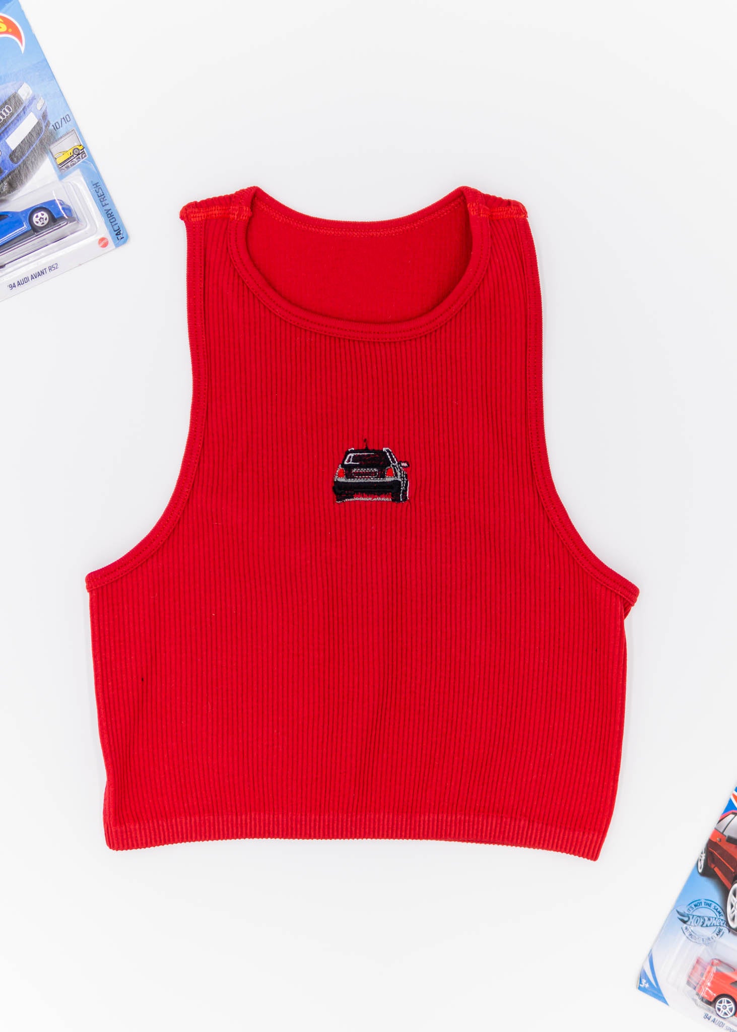A red Audi crop top for women. Photo is a front view of the top with an embroidered black Audi B5 RS4. Fabric composition is polyester, and elastine. The material is stretchy, ribbed, and non-transparent. The style of this shirt is sleeveless, with a crewneck neckline.