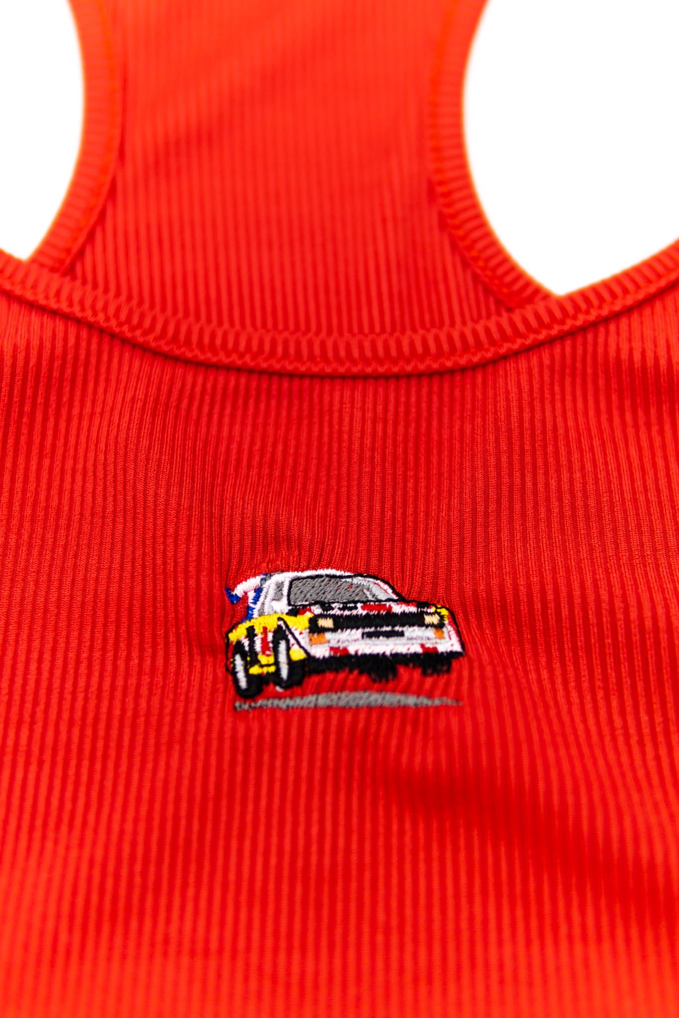 A red Audi crop top for women. Photo is a close up view of the top with an embroidered Audi Sport Quattro S1 E2. Fabric composition is polyester, and cotton. The material is stretchy, ribbed, and non-transparent. The style of this shirt is sleeveless, with a scoop neckline.