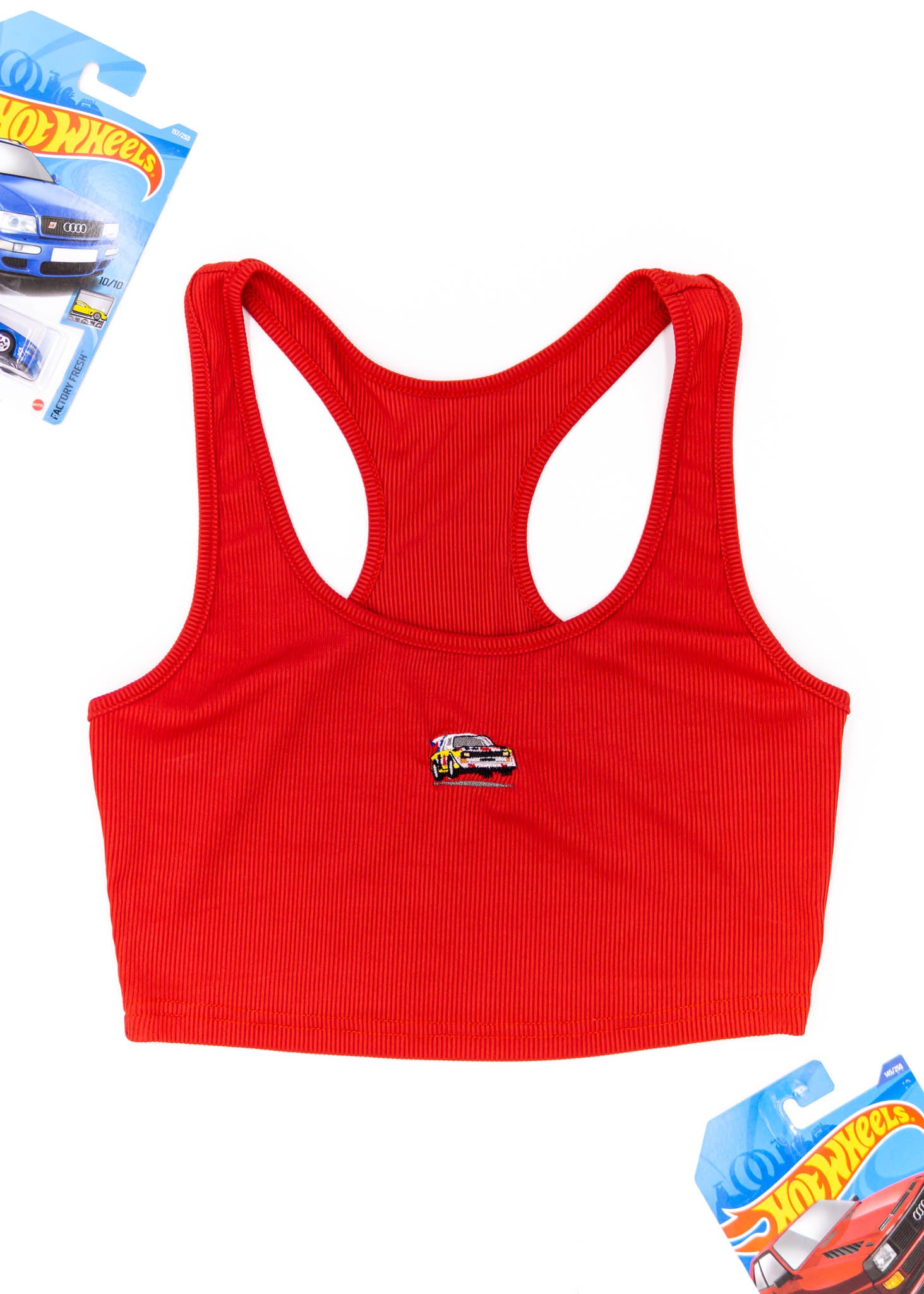 A red Audi crop top for women. Photo is a front view of the top with an embroidered Audi Sport Quattro S1 E2. Fabric composition is polyester, and cotton. The material is stretchy, ribbed, and non-transparent. The style of this shirt is sleeveless, with a scoop neckline.