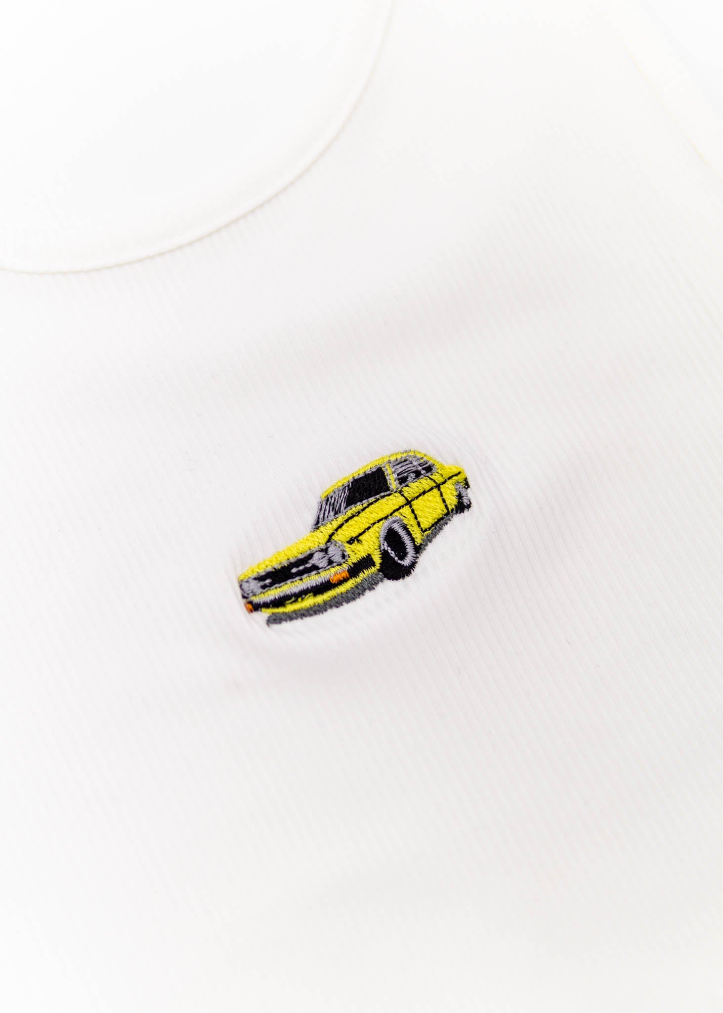 A white Audi crop top for women. Photo is a close up front view of the top with an embroidered yellow Audi 100LS. Fabric composition is polyester, and elastine. The material is stretchy, ribbed, and non-transparent. The style of this shirt is sleeveless, with a round neckline.