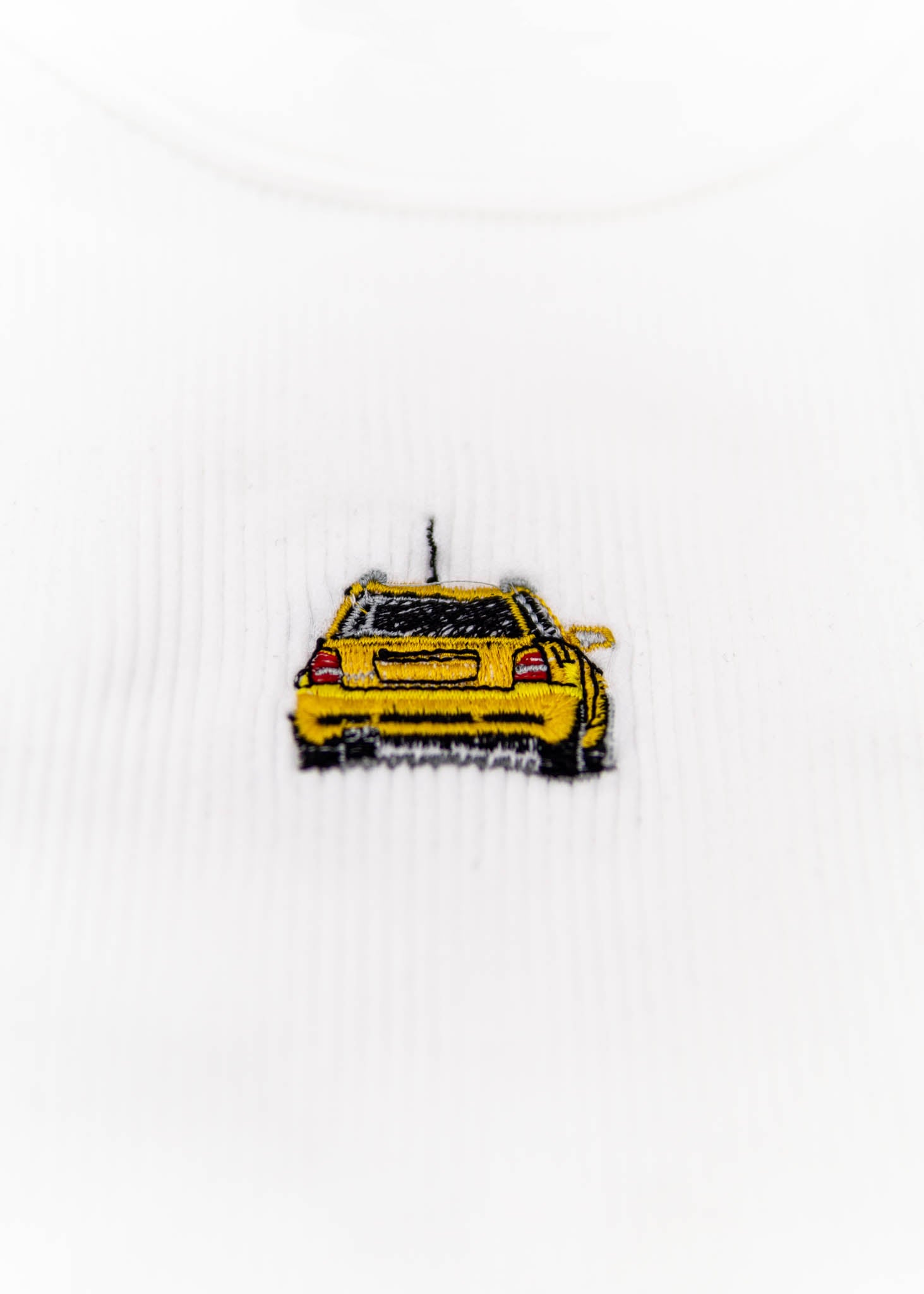 A white Audi crop top for women. Photo is a close up view of the top with an embroidered Imola Yellow Audi B5 RS4. Fabric composition is polyester, and elastine. The material is stretchy, ribbed, and non-transparent. The style of this shirt is sleeveless, with a crewneck neckline.