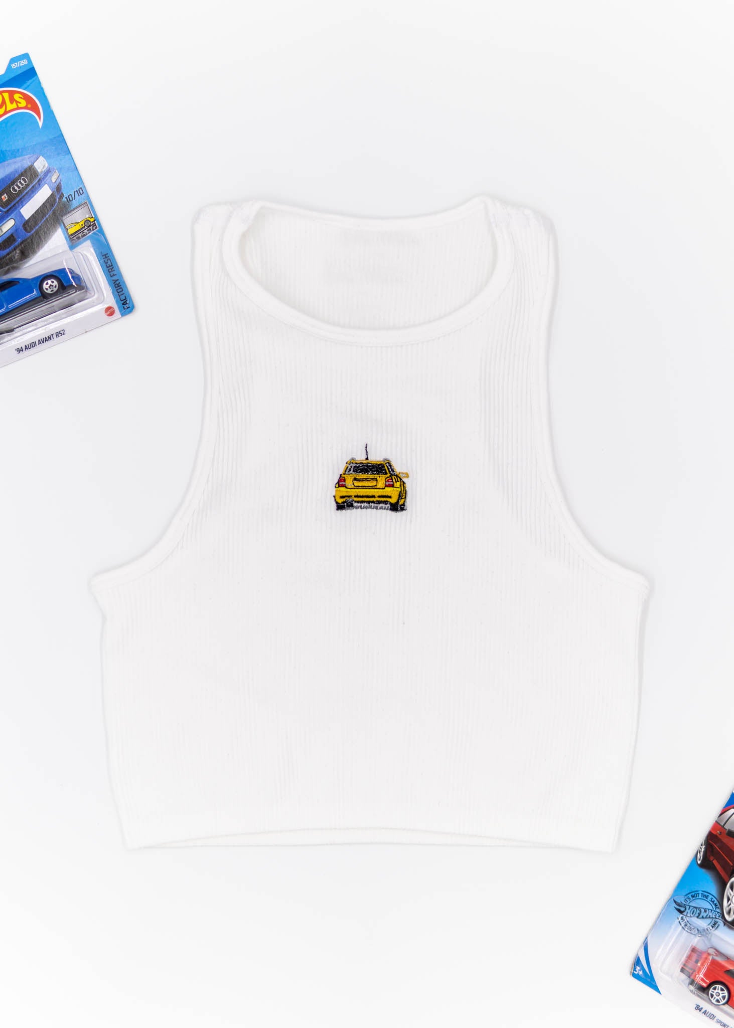 A white Audi crop top for women. Photo is a front view of the top with an embroidered Imola Yellow Audi B5 RS4. Fabric composition is polyester, and elastine. The material is stretchy, ribbed, and non-transparent. The style of this shirt is sleeveless, with a crewneck neckline.