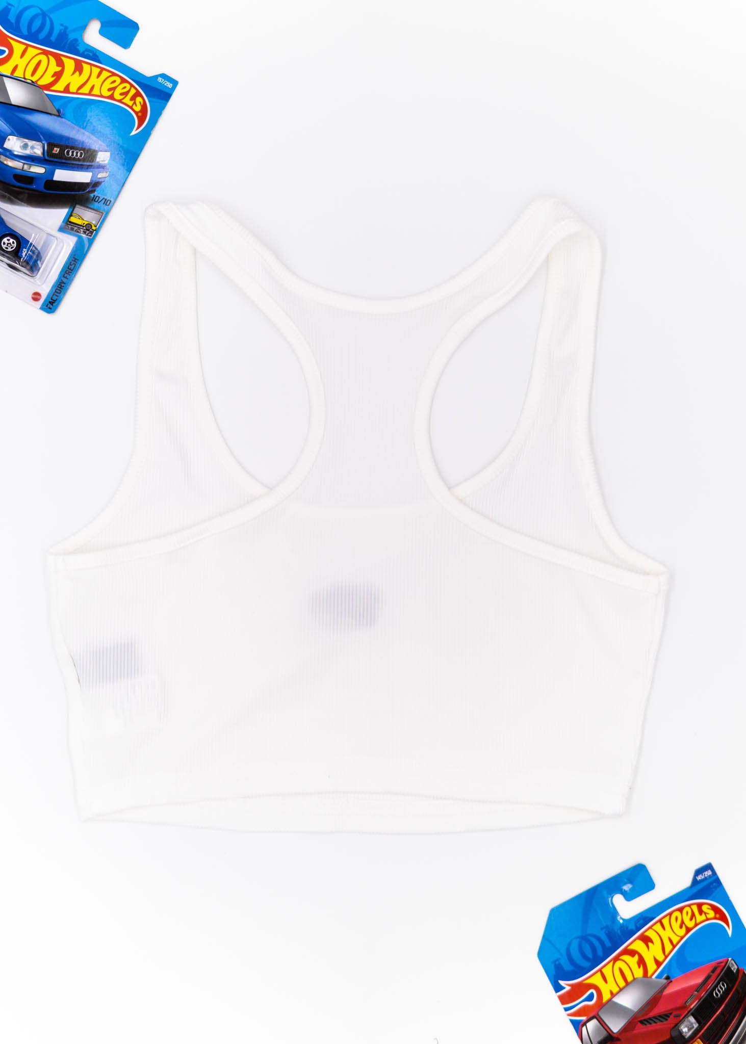 A white Audi crop top for women. Photo is a back view of the top with an embroidered Audi Sport Quattro S1 E2. Fabric composition is polyester, and cotton. The material is stretchy, ribbed, and non-transparent. The style of this shirt is sleeveless, with a scoop neckline.