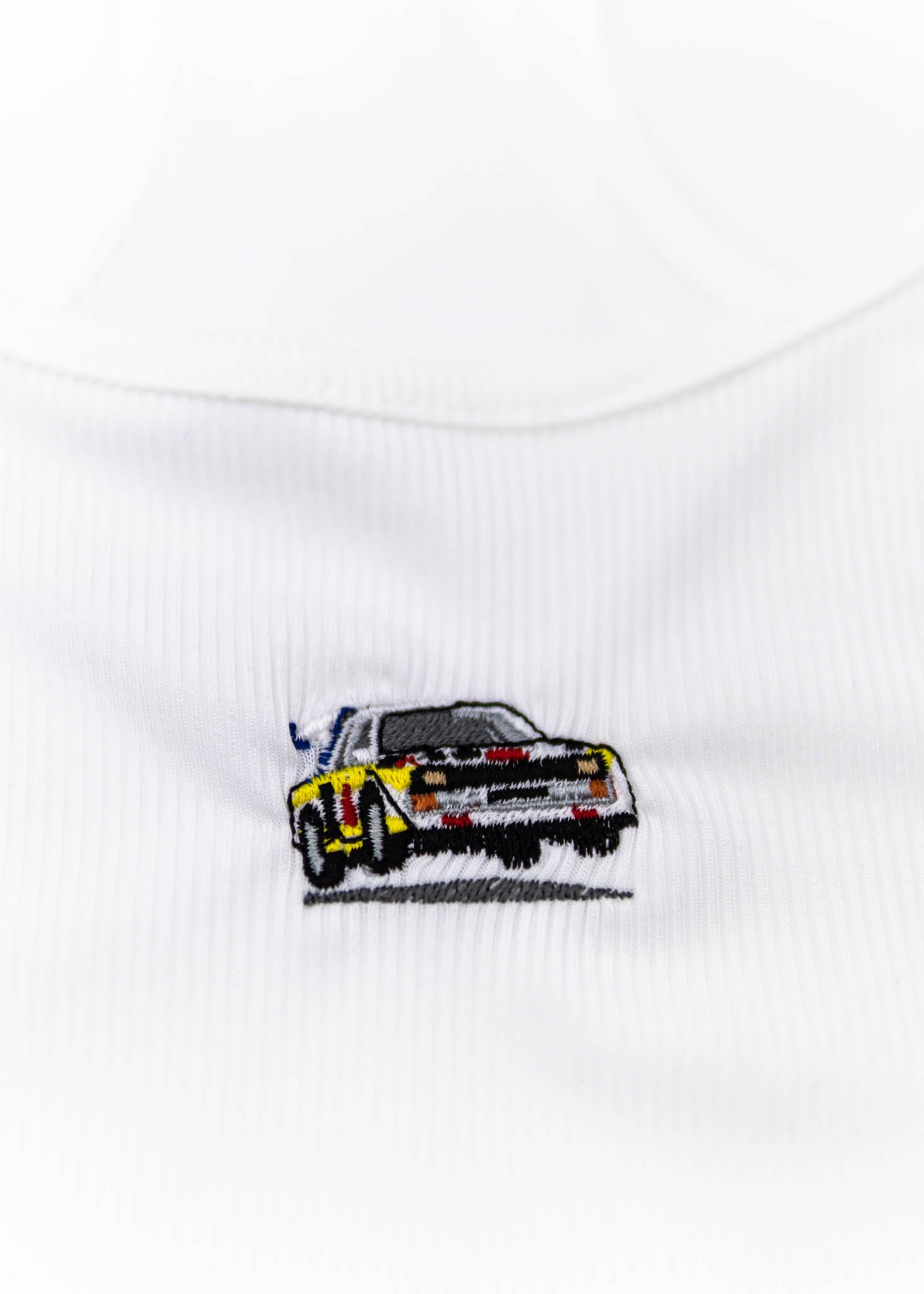 A white Audi crop top for women. Photo is a close up view of the top with an embroidered Audi Sport Quattro S1 E2. Fabric composition is polyester, and cotton. The material is stretchy, ribbed, and non-transparent. The style of this shirt is sleeveless, with a scoop neckline.
