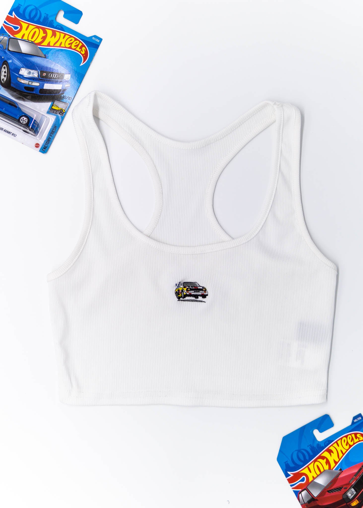 A white Audi crop top for women. Photo is a front view of the top with an embroidered Audi Sport Quattro S1 E2. Fabric composition is polyester, and cotton. The material is stretchy, ribbed, and non-transparent. The style of this shirt is sleeveless, with a scoop neckline.