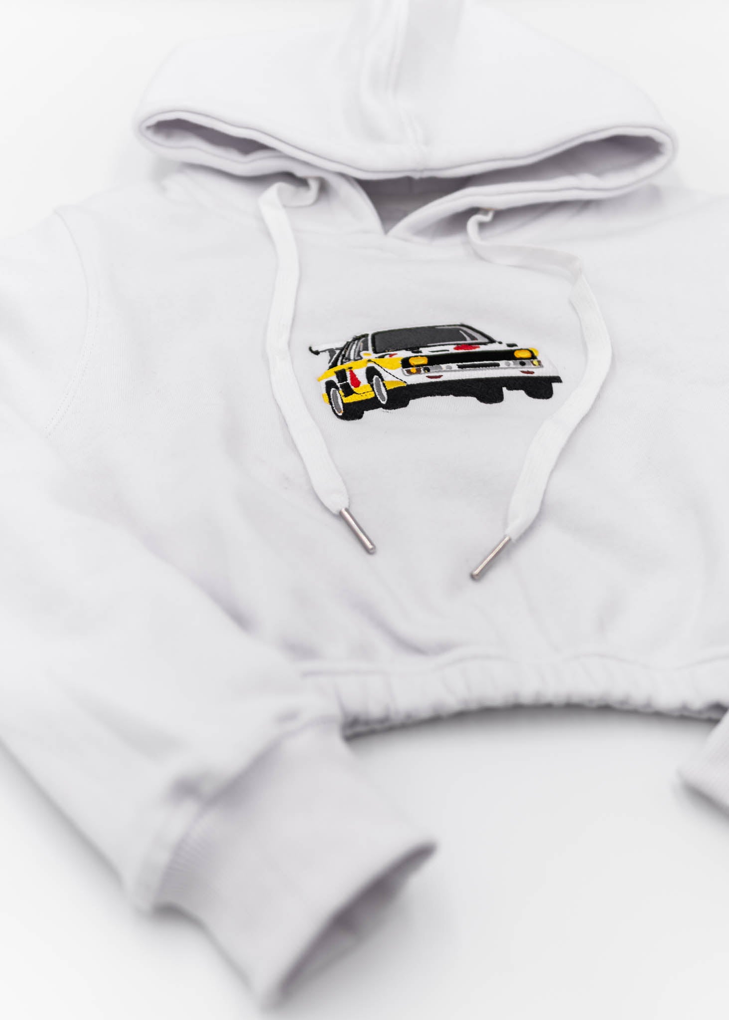 A white Audi cropped hoodie for women. Photo is a front view of the cropped sweater with an embroidered Sport Quattro S1 E2. Fabric composition is cotton, and polyester. The material is soft, stretchy, and non-transparent. The style of this crop hoodie is long sleeve, crewneck with a hood, hooded, with embroidery on the chest.